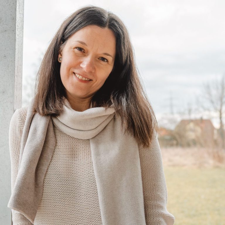 Simona Singer, Kita- und Familienfotografin Frau mit langen, dunklen Haaren, lächelt in bequemer Kleidung vor einer Landschaft.